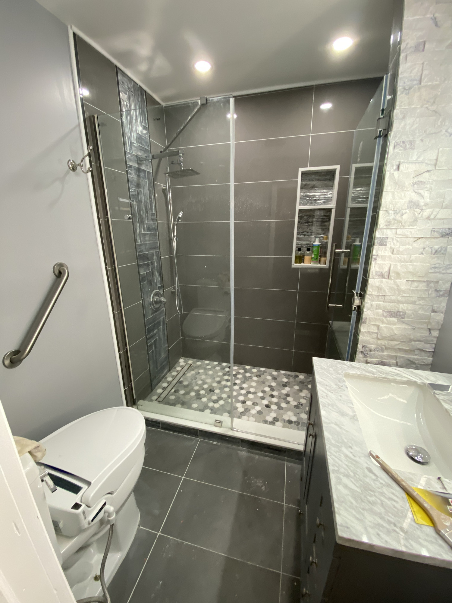 Gray tiled shower with hex-mosaic floor, recessed shelves, glass enclosure, and handheld fixture