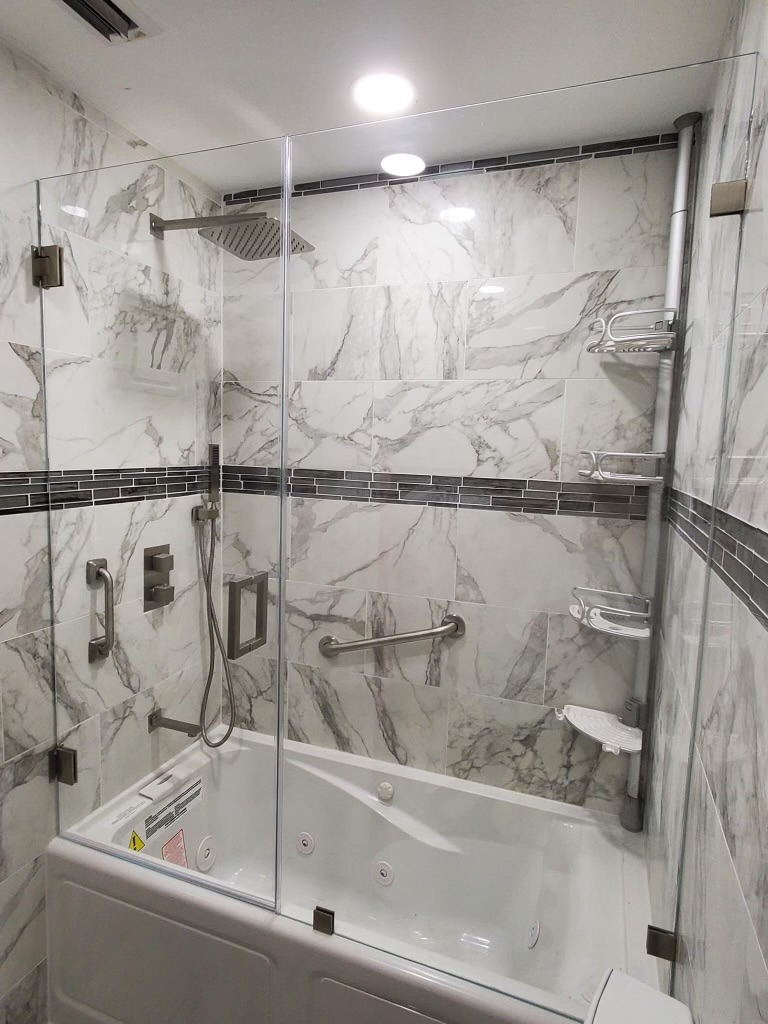 Marble-look tiled shower enclosure with dark accent band, handheld fixture, glass, and grab bars